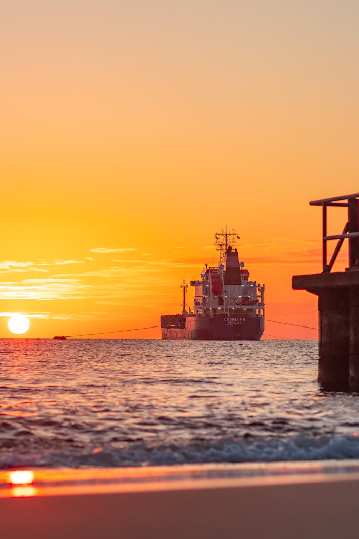 A cargo ship sails on the ocean during a stunning sunset, emitting a warm glow.